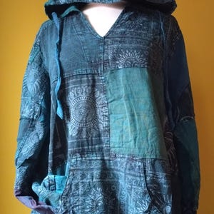 Op de afbeelding: Een teal en blauwe patchwork hoodie met een capuchon en een voorzak. De hoodie heeft een V-hals en is gemaakt van stof met patronen met zon- en bloemmotieven. Het kledingstuk heeft een relaxte pasvorm en lange mouwen.