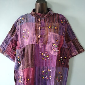 May include: A patchwork shirt in shades of purple, brown, and blue. The short-sleeved shirt features a mandarin collar, wooden buttons, and a chest pocket. The fabric is decorated with embroidered floral designs.