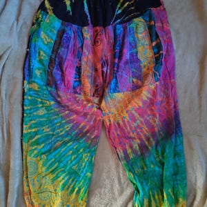 Op de afbeelding: Veelkleurige tie-dye harembroek met een elastische tailleband. De kleuren zijn onder meer blauw, groen, geel, oranje, paars en roze. De stof heeft een wervelend tie-dye patroon.