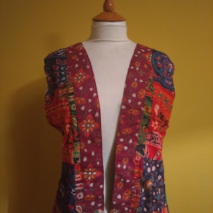 May include: A sleeveless vest featuring a patchwork design with various floral and geometric patterns in shades of deep red, orange, blue, and green. The garment has a wide, solid-colored maroon border along the front opening with a floral print.