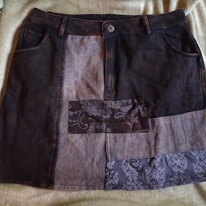 May include: A dark brown denim skirt with a patchwork design. The skirt features a button closure, pockets, and panels of grey and patterned fabric. The skirt's design includes floral and damask patterns.