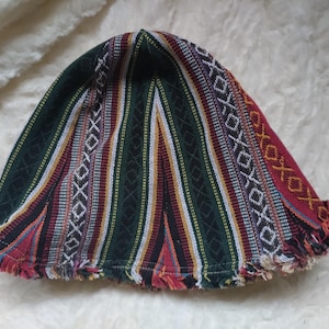 May include: A patterned hat with a brim and fringe. The hat features vertical stripes in green, red, yellow, and white, with a geometric design. The brim is trimmed with a fringe.