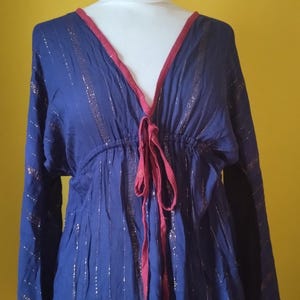 May include: A long-sleeved blue tunic featuring vertical metallic gold stripes throughout the fabric. The garment has a deep V-neckline trimmed in dark pink, with a matching drawstring tie at the centre of the chest and an elasticated waist.