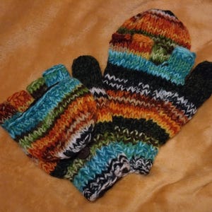 Könnte beinhalten: Ein Paar handgestrickte Fäustlinge und fingerlose Handschuhe. Die Fäustlinge und Handschuhe sind mehrfarbig mit Streifen in Blau, Orange, Grün, Schwarz und Weiß. Die fingerlosen Handschuhe haben einen Knopfverschluss. Der Hintergrund ist ein warmer, brauner Farbton.