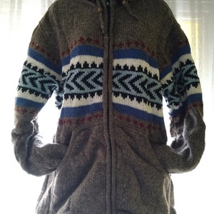 May include: A grey, hooded zip-up jacket with a geometric pattern in blue, white, and black across the chest. The jacket has a drawstring at the bottom and two front pockets. It is made of a thick, textured material, suitable for cooler weather.