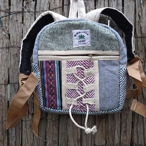 Puede incluir: Una mochila de cáñamo con diseño patchwork. La bolsa presenta una mezcla de telas azules, moradas y crema. Tiene correas ajustables color canela, un bolsillo con cremallera y un panel frontal con un patrón tejido decorativo y un logotipo de hoja de cáñamo.