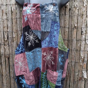 May include: Patchwork dungaree dress in shades of blue, green, purple, and black. The dress features embroidered floral designs and decorative stitching. The straps are a matching grey velvet material. The dress is displayed on a black mannequin.