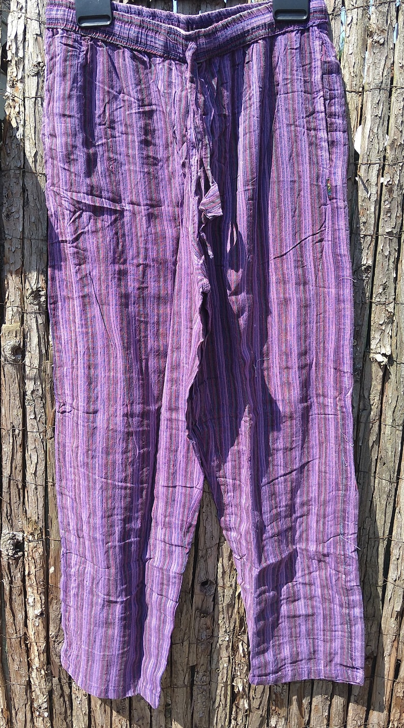 May include: Purple striped pants with a drawstring waist. The pants have vertical stripes in shades of purple, lavender, and brown. The fabric appears to be lightweight and suitable for casual wear. The pants are hanging against a wooden background.