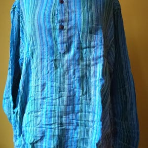 Peut inclure: Une chemise à manches longues rayée verticalement en nuances de bleu et de vert. La chemise a un col mandarin, une petite poche et trois boutons marron. Le tissu semble léger et texturé.