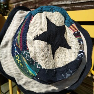 Peut inclure: Chapeau seau beige avec une étoile noire. Le chapeau présente un motif patchwork avec des tissus bleus, verts, rouges et noirs. Le bord est noir. Le diamètre du chapeau est d'environ 25 cm.