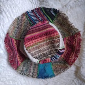 May include: A patchwork bucket hat with a colourful, fringed brim. The hat features a variety of patterned fabrics in shades of red, pink, blue, green, and brown. The hat's crown is a lighter colour, and the brim is made of a woven material.