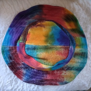 Op de afbeelding: Een bucket hat met een levendig tie-dye patroon in gemengde kleuren zoals paars, blauw, groen, geel, oranje en rood. De hoed heeft een ronde kroon en een brede, naar beneden lopende rand met zichtbare witte, cirkelvormige stiksels over het geheel.