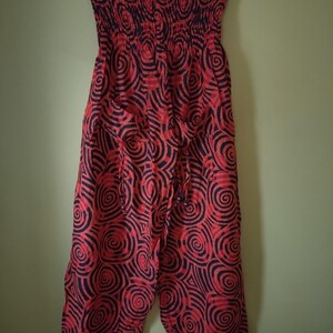 May include: A strapless jumpsuit featuring a vibrant red and black spiral pattern throughout. The garment has a smocked, elasticized bodice and loose, wide-leg trousers that gather at the ankles. Two front pockets are visible on the hips.