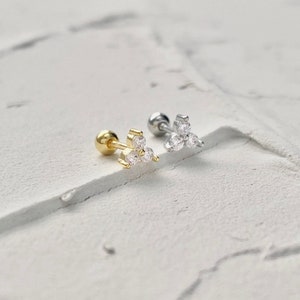 May include: Two small triangle shaped earrings with three clear cubic zirconia stones each. The earrings are on a white textured surface. One earring is gold and the other is silver.