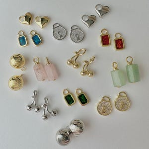 May include: A collection of jewelry charms including gold and silver heart shapes, padlock pendants with clear stones, rectangular charms with blue, red, and green inserts, pink and green stone cylinders, gold and silver cherry charms, and spheres.