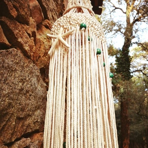 Peut inclure: Une lampe suspendue en macramé blanche avec des perles vertes et une décoration en forme d'étoile de mer. La lampe est suspendue à un mur en pierre.