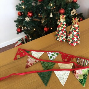 May include: A red and green Christmas banner with gold accents hangs on a wooden table. Two small, handmade Christmas trees made of fabric are on the table. The trees are decorated with red, green, and gold fabric.