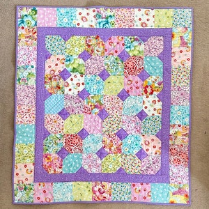 Könnte beinhalten: Eine lila und weiße Patchwork-Decke mit Blumenmuster. Die Decke besteht aus vielen kleinen Stoffquadraten, die jeweils ein anderes Blumenmuster haben. Die Decke ist mit einem lilafarbenen Rand eingefasst.