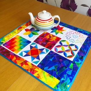 Può includere: Un runner da tavolo in patchwork colorato con un design arcobaleno. Il runner è fatto di quadrati di tessuto con diversi motivi geometrici. Un bollitore bianco a righe arcobaleno si trova sopra il runner.