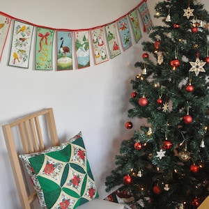 May include: A green and red quilted pillow sits on a wooden chair in front of a decorated Christmas tree. A red and white string of Christmas flags with numbers and illustrations hangs on the wall behind the chair.