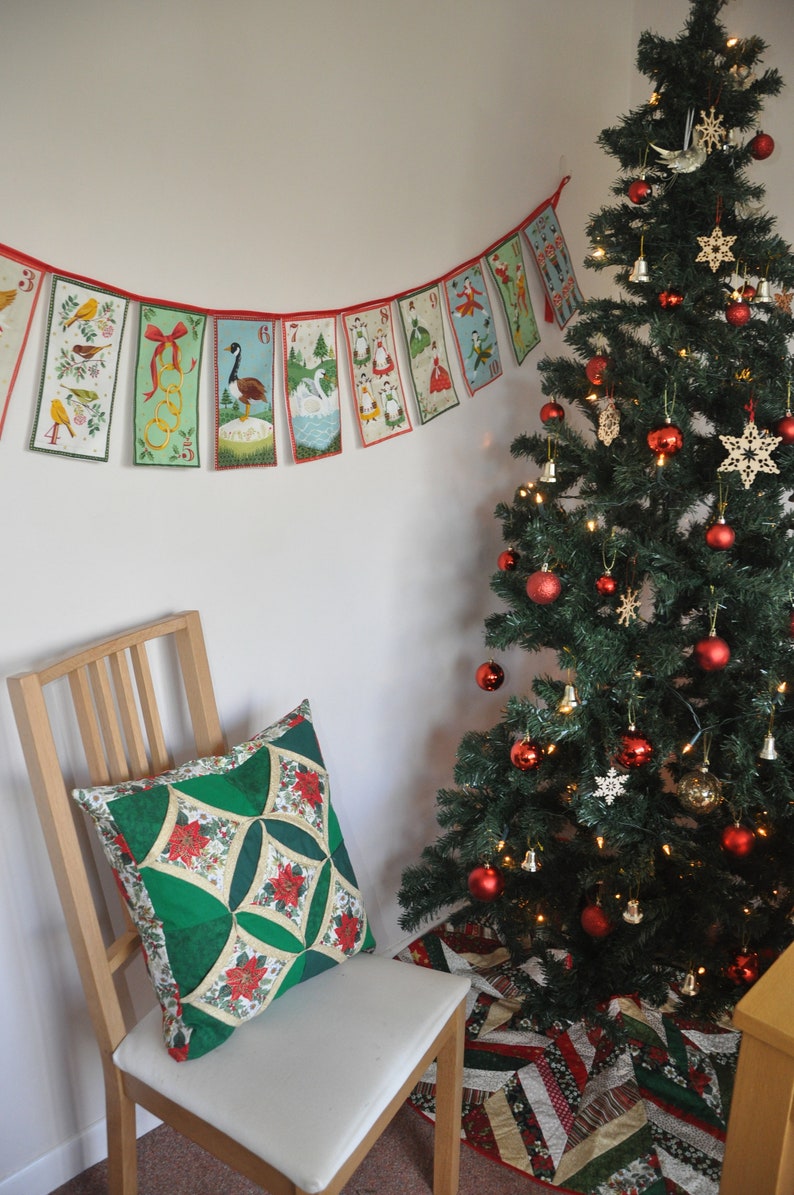 May include: A green and red quilted pillow sits on a wooden chair in front of a decorated Christmas tree. A red and white string of Christmas flags with numbers and illustrations hangs on the wall behind the chair.