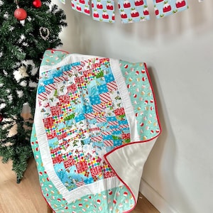 May include: A patchwork quilt with a Christmas theme. The quilt is made of various fabrics with different Christmas patterns, including candy canes, presents, and Christmas trees. The quilt is folded and draped over a wooden chair. A Christmas garland with red and white Christmas pudding designs hangs on the wall behind the chair.