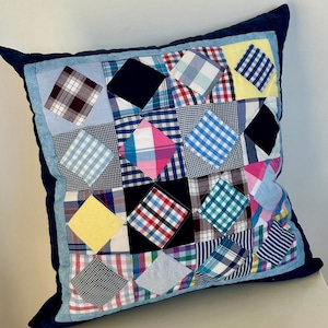 May include: A blue and white patchwork pillow with a navy blue border. The pillow is made of different plaid fabrics in various shades of blue, yellow, pink, and black.