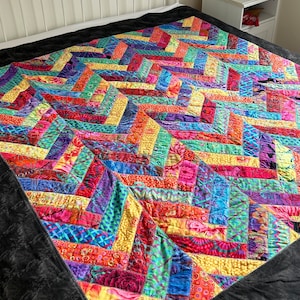 Op de afbeelding: Een kleurrijke, handgemaakte quilt met een patchwork-ontwerp. De quilt heeft een verscheidenheid aan stoffen in tinten rood, oranje, geel, groen, blauw en paars. Het heeft een zwarte rand en wordt op een bed getoond. De quilt meet ongeveer 203 cm bij 203 cm.