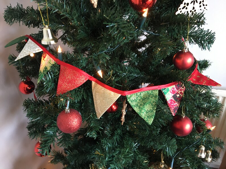 May include: A red, green, and gold Christmas bunting with a red ribbon, hanging on a Christmas tree decorated with red ornaments and lights.