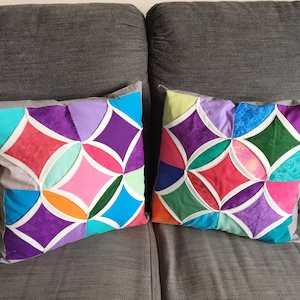 May include: Two colorful geometric patterned throw pillows with white piping on a gray couch. The pillows are made of different fabrics and feature a variety of colors, including purple, pink, blue, green, and orange.
