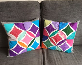Puede incluir: Dos cojines de sofá geométricos coloridos con ribete blanco sobre un sofá gris. Los cojines están hechos de diferentes telas y presentan una variedad de colores, incluyendo morado, rosa, azul, verde y naranja.