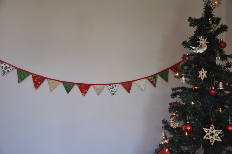 May include: A red, green, and gold Christmas banner with a festive pattern hangs on a white wall. A decorated Christmas tree with red ornaments, wooden stars, and a silver bird stands to the right.