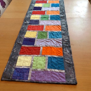 May include: A colorful table runner with a patchwork design featuring squares of various colors, including red, orange, yellow, green, blue, purple, and gray. The runner has a gray border with a subtle sparkle.