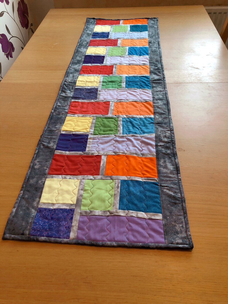 May include: A colorful table runner with a patchwork design featuring squares of various colors, including red, orange, yellow, green, blue, purple, and gray. The runner has a gray border with a subtle sparkle.