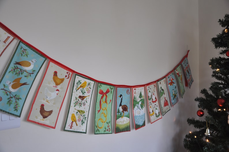 May include: A Christmas advent calendar banner with red and white string and 12 flags. Each flag has a different number, animal, and design. The flags are in a curved shape.