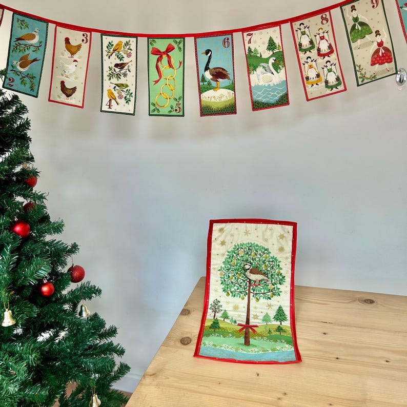 May include: A Christmas advent calendar banner with red and white numbers, featuring images of birds, swans, and angels. The banner is hanging on a white wall, and a small quilted Christmas tree with a bird on it is on a wooden table.