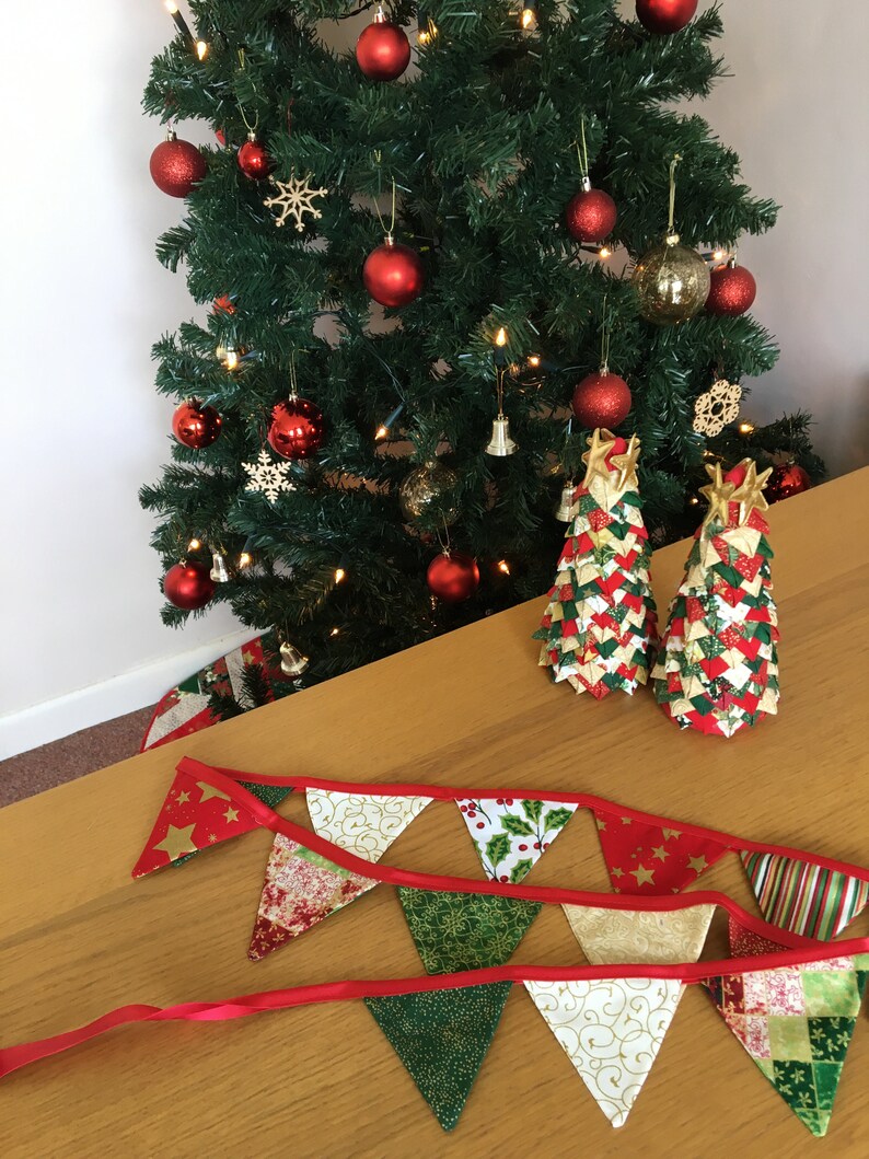May include: A red and green Christmas banner with gold accents hangs on a wooden table. Two small, handmade Christmas trees made of fabric are on the table. The trees are decorated with red, green, and gold fabric.