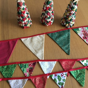 May include: A festive Christmas bunting with red, green, and white fabric triangles. The bunting has a red ribbon and is decorated with gold glitter and various patterns. Three small Christmas trees made of fabric are in the background.