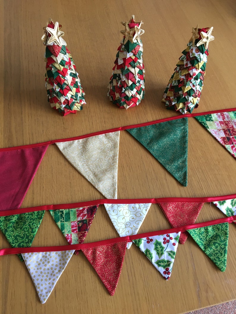 May include: A festive Christmas bunting with red, green, and white fabric triangles. The bunting has a red ribbon and is decorated with gold glitter and various patterns. Three small Christmas trees made of fabric are in the background.