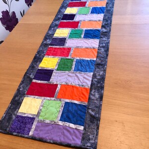 May include: A colorful table runner with a rainbow pattern of squares framed in white. The runner has a gray border with a subtle sparkle.