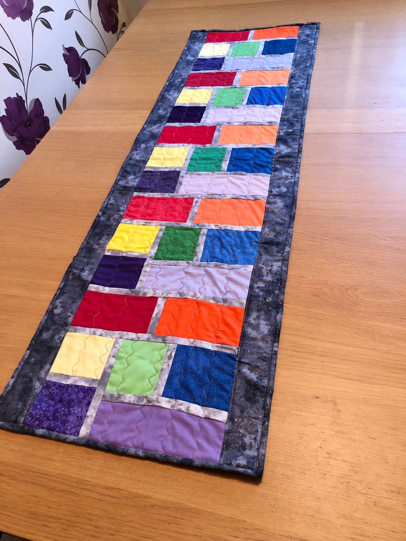 May include: A colorful table runner with a rainbow pattern of squares framed in white. The runner has a gray border with a subtle sparkle.