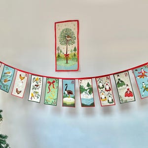 May include: A red and white Christmas advent calendar banner with a variety of illustrations, including birds, a swan, a goose, and dancing figures. The banner is hung on a white wall with a Christmas tree in the foreground.