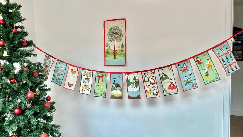May include: A red and white Christmas advent calendar banner with a variety of illustrations, including birds, a swan, a goose, and dancing figures. The banner is hung on a white wall with a Christmas tree in the foreground.