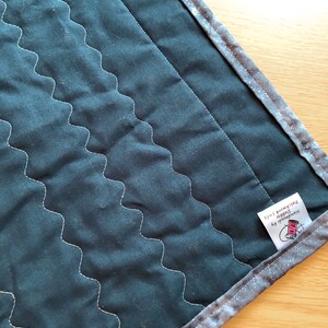 May include: A dark blue quilted fabric with a white zig-zag stitch pattern. The fabric has a silver and gray border with a white label that reads "Handmade by Debbie Patchwork Lady".
