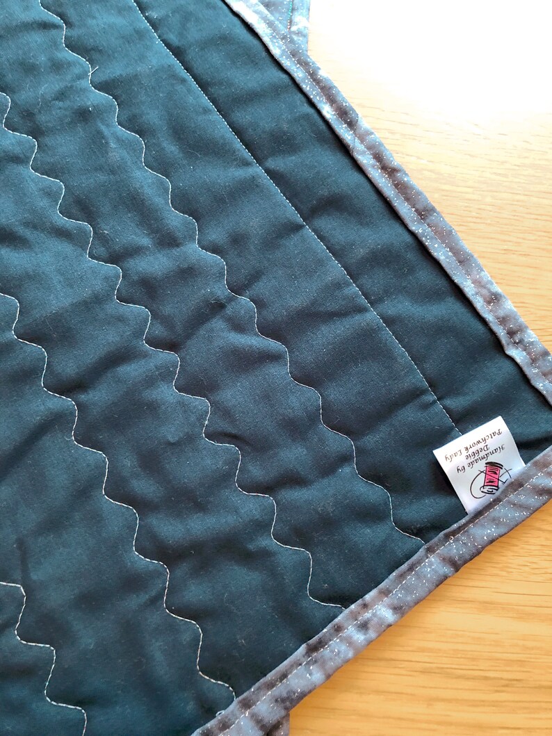 May include: A dark blue quilted fabric with a white zig-zag stitch pattern. The fabric has a silver and gray border with a white label that reads "Handmade by Debbie Patchwork Lady".