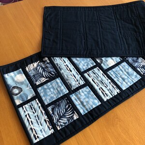 May include: Two quilted table runners, one navy blue and one with a patchwork design of blue and white geometric patterns. The patchwork runner has a navy blue border.