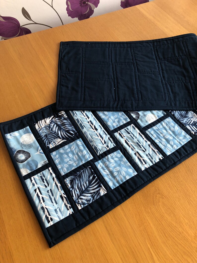 May include: Two quilted table runners, one navy blue and one with a patchwork design of blue and white geometric patterns. The patchwork runner has a navy blue border.