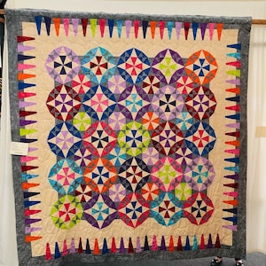 Puede incluir: Una colcha colorida con un patrón geométrico de círculos y triángulos. La colcha está hecha con una variedad de telas en tonos de rosa, morado, azul, verde, rojo y naranja. La colcha está enmarcada con un borde gris y una fila de triángulos coloridos.