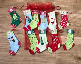 May include: A set of 12 small Christmas stockings with various designs, including a Christmas tree, Santa Claus, and an elf. The stockings are red, green, and blue with white polka dots. The numbers 1 through 12 are printed on the stockings.