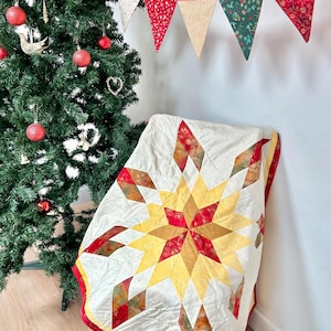 May include: A white quilt with a red, yellow and orange starburst pattern. The quilt is folded and draped over a chair. A Christmas tree with red and gold baubles is in the background.